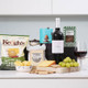 Wine & Gourmet Delights Hamper. A neatly arranged tabletop featuring a variety of snacks and beverages. On the left, a bag of Keogh's Mature Irish Cheese and Onion crisps, alongside a wooden board with crackers, cheese slices, and green grapes. A bottle of red wine stands centrally, with a glass filled beside it. A box of Forest Feast almonds is visible in the background, along with a decorative container for snacks. The setting emphasizes a casual dining or tasting experience. Wine & Gourmet Delights Hamper. A neatly arranged tabletop featuring a variety of snacks and beverages. On the left, a bag of Keogh's Mature Irish Cheese and Onion crisps, alongside a wooden board with crackers, cheese slices, and green grapes. A bottle of red wine stands centrally, with a glass filled beside it. A box of Forest Feast almonds is visible in the background, along with a decorative container for snacks. The setting emphasizes a casual dining or tasting experience.